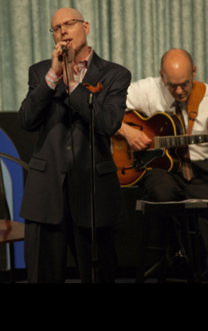crooning musician clutching microphone with guitar player strumming behind him.
